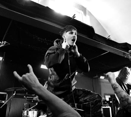 Ung mand, der står på en scene med fingrene til ørene, mens publikum laver metal horn